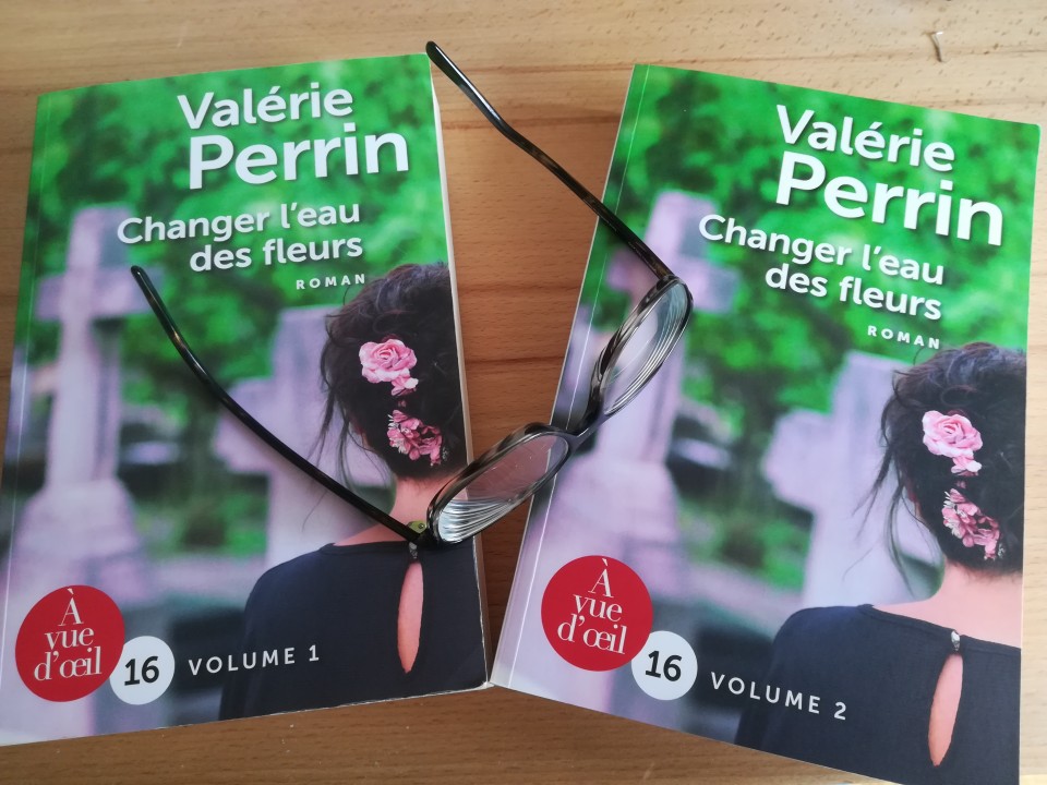 Posés sur une table en bois de chêne, les deux volumes du roman "Changer l'eau des fleurs" de Valérie Perrin, sur lesquelles est posée, de travers, une paire de lunettes de vue, branches déployées vers la gauche de la photo. On devine que ce sont des doubles-foyers.